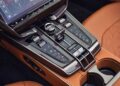Close-up of the 2026 Aston Martin Vanquish’s center console featuring a touchscreen display, control knobs, buttons, gear selector, cup holders, and brown leather upholstery—highlighting its luxurious V12 craftsmanship.