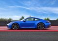 A Limited-Run 2026 911 Carrera T Club Coupe, celebrating 70 years of Porsche Club of America, is parked on a red and white striped surface with a black barrier and trees in the background under a blue sky.