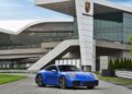 A blue Limited-Run 2026 911 Carrera T Club Coupe is parked in front of a modern glass building with a large Porsche logo on the facade, celebrating 70 years of Porsche Club of America.