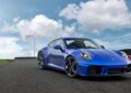 A blue Limited-Run 2026 911 Carrera T Club Coupe is parked on a racetrack under a partly cloudy sky, celebrating 70 Years of Porsche Club of America with a flag waving in the background.