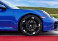 Close-up side view of the Limited-Run 2026 911 Carrera T Club Coupe, highlighting its front wheel and bumper on a red track, with green grass and blue sky—celebrating 70 years of the Porsche Club of America.