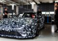 A Porsche 911 Cup Car with black and white camouflage paint is parked in a garage as four people stand nearby, two on each side, preparing the car for the upcoming 2026 Race Season in a workshop setting.