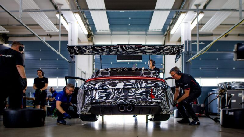 A next-gen Porsche 911 Cup Car with a camouflage wrap is serviced by a pit crew inside a garage, surrounded by equipment and tools, ready for the 2026 race season.