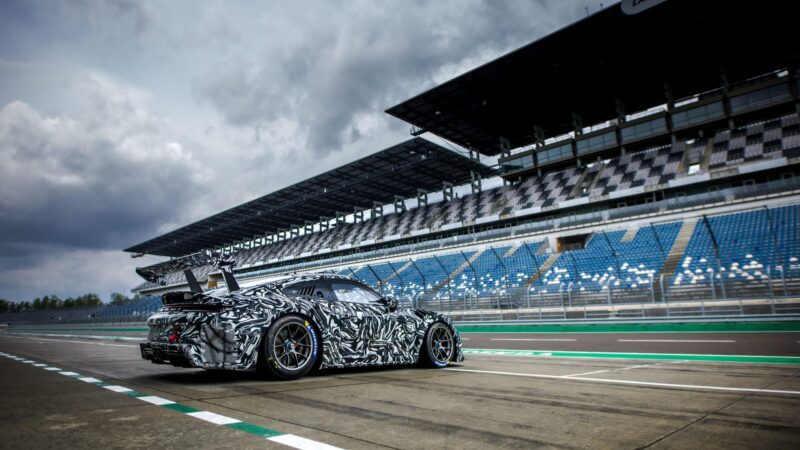 A Porsche 911 Cup Car with a camouflage wrap is parked on a racetrack in front of an empty grandstand under a cloudy sky, ready for the 2026 Race Season.