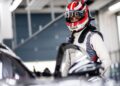 A race car driver in a Porsche-branded helmet and racing suit stands next to a Porsche 911 Cup car in a garage, preparing for the next-gen 2026 race season.
