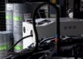 Three barrels labeled “eFuels Porsche” stand beside industrial equipment and a Porsche 911 Cup car part, hinting at next-gen technology for the 2026 race season inside a modern garage or workshop.