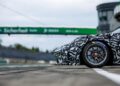 A camouflaged next-gen Porsche 911 Cup race car is parked on a track, showing its front wheel and partial side view, with a blurred raceway and bridge in the background, hinting at what's coming for the 2026 race season.