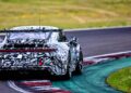 A camouflaged next-gen Porsche 911 Cup Car with a rear wing drives on a racetrack, rounding a corner bordered by grass and painted curbs, hinting at the excitement of the 2026 race season.