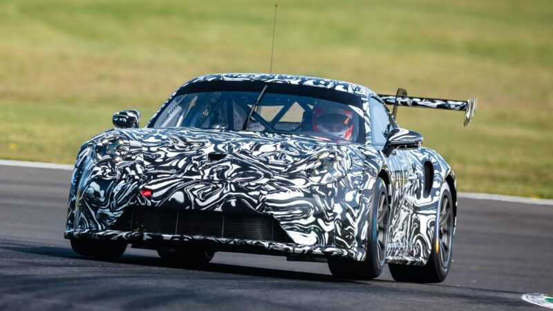 A next-gen Porsche 911 Cup Car with black and white camouflage wrap speeds on a racetrack, blurred grass in the background—hinting at its debut for the 2026 race season.