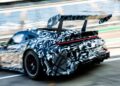 A black and white camouflaged Porsche 911 Cup Car with a large rear wing speeds along the racetrack, passing empty stadium seats, signaling excitement for the 2026 Race Season.