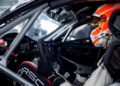 A race car driver wearing a helmet and racing suit sits in the cockpit of a Next-Gen Porsche 911 Cup Car, hands on the steering wheel, surrounded by safety bars and controls, ready for the 2026 Race Season.