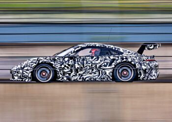 A next-gen race car with black-and-white camouflage wraps speeds along a track, motion blur highlighting its pace, and a large rear wing visible—hinting at innovations for the 2026 race season.