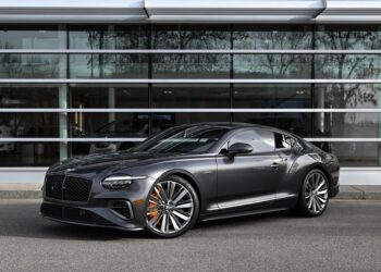 A dark gray luxury coupe, one of the best Bentleys with large alloy wheels, is parked in front of a modern glass building.