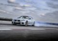 A silver 2025 BMW coupe drifts on a racetrack at Monterey Car Week, producing tire smoke against a cloudy sky.
