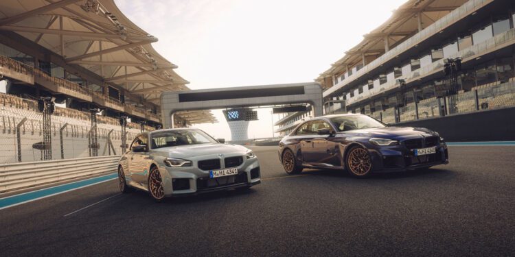 Two BMW cars are parked side by side on a racetrack at Monterey Car Week 2025, with empty grandstands and a modern structure in the background under a bright sky.