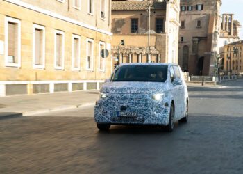 A camouflaged Mercedes-Benz prototype, possibly the VLE Electric Van, with a German license plate is being tested on a city street in Rome, with historic buildings in the background.