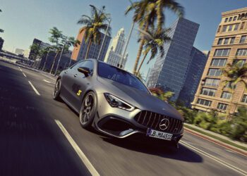 A matte grey Mercedes-Benz sports car drives down a palm-lined city street with modern buildings in the background under a clear sky.