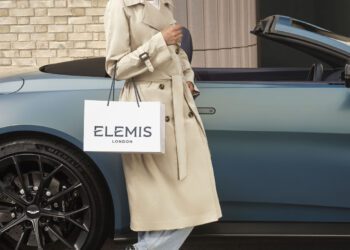 Person in a beige trench coat stands beside a blue convertible car, holding an Elemis London shopping bag.
