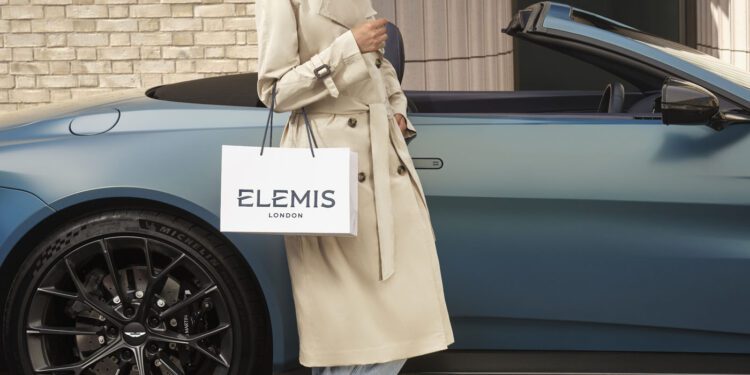 Person in a beige trench coat stands beside a blue convertible car, holding an Elemis London shopping bag.