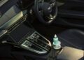 A bottle of car cleaner, a microfiber cloth, and a pair of gloves are placed on the center console of a modern car with leather seats and a digital dashboard.