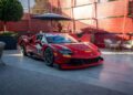 A red Ferrari race car with the number 7 on the door is parked on a concrete patio with outdoor furniture and red paneling in the background, reminiscent of a scene at Goodwood.