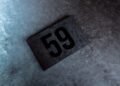 A rectangular black plaque with the number 59 in large, bold black font rests on a textured gray concrete surface, reminiscent of the legendary McLaren F1 GTR that triumphed at Goodwood with Lanzante.