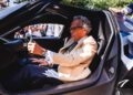 An older man wearing a beige jacket and glasses sits inside a black Zenvo Aurora sports car, gripping the steering wheel, with a crowd standing outside during its global debut.