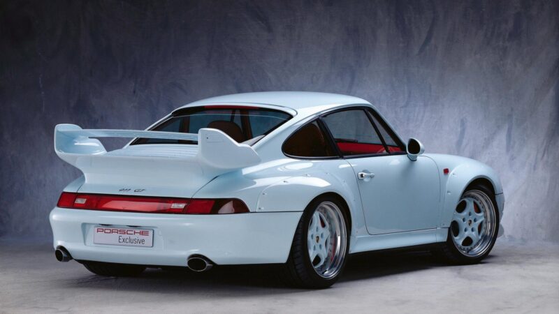 A white Porsche 911 GT sports car is shown from the rear at an angle, featuring a large rear wing and Porsche Exclusive branding on the license plate—highlighting Porsche exclusive requests for factory one-off cars.