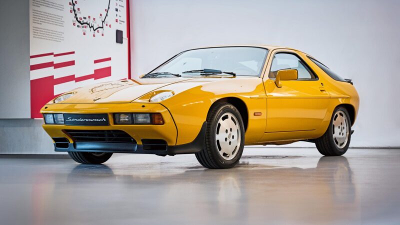 A yellow vintage sports car is displayed indoors on a shiny floor, with informational panels in the background—an example of Porsche exclusive requests and factory one-off cars.