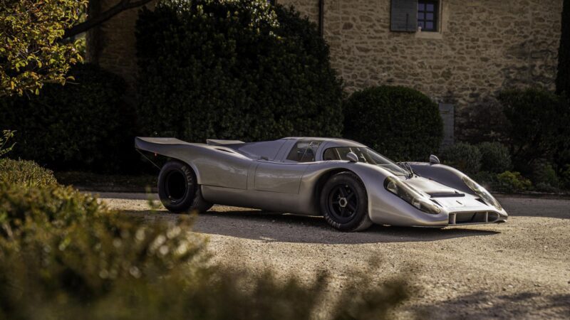 A vintage silver Porsche race car with a low, aerodynamic body—possibly one of the remarkable factory one-off cars—is parked on a gravel driveway in front of a stone building and lush greenery.