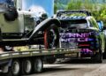 A modern SUV transports a vintage Porsche 911 on a flatbed trailer along a road bordered by greenery and a metal fence, reminiscent of something Richard Hammond might admire.