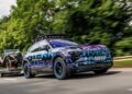 A Porsche SUV with a colorful camouflage wrap, possibly inspired by Richard Hammond, tows a vintage Porsche 911 on a trailer, cruising down a road with blurred greenery in the background.
