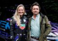 Two people stand smiling in front of a Porsche 911 race car; one, possibly Richard Hammond, wears a racing suit with sponsor logos, while the other is dressed in casual clothing.