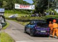 A Porsche 911 with a colorful wrap is at the starting line of the Shelsley Walsh hill climb event, with officials in orange suits nearby, spectators watching, and Richard Hammond rumored to be among the attendees.