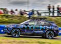 A Porsche 911 SUV with blue and purple camouflage wrap, reminiscent of Richard Hammond's style, speeds along a race track with blurred spectators and greenery in the background.
