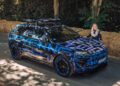 A woman stands next to a camouflaged Porsche SUV with blue and black graphics, parked beside a road lined with hay bales and trees, evoking the spirit of GT3 race cars.