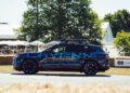 A camouflage-wrapped Porsche SUV prototype, possibly hinting at GT3-inspired features, is driving on a road at an outdoor event, with trees and tents visible in the background.