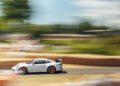 A white Porsche sports car speeds along the Goodwood racetrack, with a blurred background indicating motion and greenery visible on a sunny day.