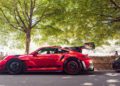 A red Porsche GT3 RS sports car is parked on a shaded road with its rear wing raised, surrounded by trees and other vehicles, showcasing the iconic GT3 performance inspired by Porsche prototypes.