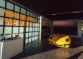 A yellow Porsche sports car is displayed indoors next to a glass case, with the words "YOU DREAM IT. WE BUILD IT." on the wall behind, capturing the spirit of innovative prototypes and race cars.