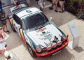 Overhead view of a white Porsche racing car with Martini livery on display outdoors, surrounded by people on a gravel surface.