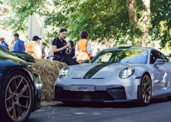 A silver Porsche 911 GT3 is parked on a shaded road at Goodwood near hay bales, with several people in the background wearing safety vests and casual clothing.