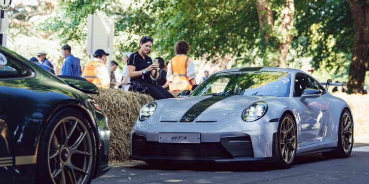 A silver Porsche 911 GT3 is parked on a shaded road at Goodwood near hay bales, with several people in the background wearing safety vests and casual clothing.
