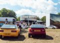 Two vintage sports cars with UK plates are parked on grass at an outdoor car show, with people walking and a Porsche GT3 display featuring prototypes in the background under a sunny sky.