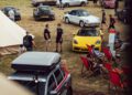 A car show on a grassy field featuring various sports cars, including a yellow Porsche convertible and a stunning GT3, with people walking, talking, and sitting in red deck chairs.