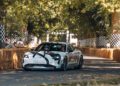 A white Porsche sports car drives on a tree-lined track bordered by straw bales and checkered flags, with spectators and safety personnel nearby. The event showcases GT3 prototypes in action.