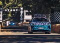 A camouflaged Porsche 911 drives down a tree-lined track toward a starting line, with hay bales and checkered flags marking the course—a scene reminiscent of a classic Richard Hammond adventure.