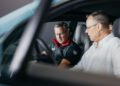 Two men sit in the front seats of a Porsche, one pointing at the dashboard as they discuss something inside the vehicle.