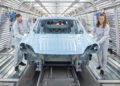 Two workers in white coveralls inspect a light blue Macan car body on an assembly line inside a brightly lit, metallic Porsche automotive manufacturing facility.