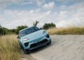 A light blue Porsche Macan drives on a steep, paved incline surrounded by tall grass, under a partly cloudy sky.